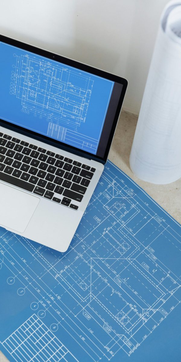 Laptop showing engineering blueprint with rolled paper and drawings on table.