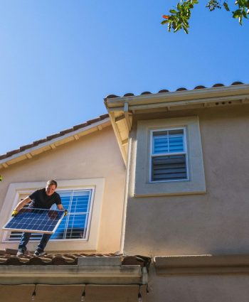 Man installing a solar panel on a rooftop under a clear blue sky, promoting renewable energy.
